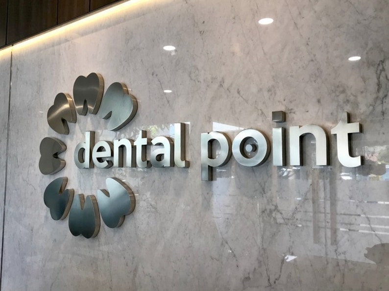 May include: A wall-mounted sign featuring the text "dental point" in metallic silver lettering. To the left of the text is a circular logo composed of six stylized tooth shapes arranged in a ring, all mounted on a white marble surface.