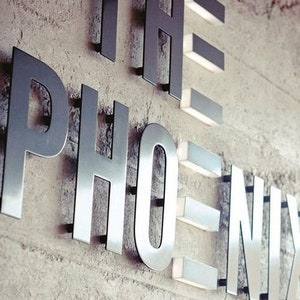 May include: The word "PHOENIX" in large, metallic letters is mounted on a textured, light-colored wall. The letters are illuminated with small, rectangular lights, creating a modern and stylish design. The sign is likely for a business or establishment.