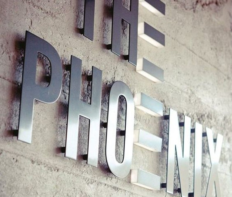 May include: The word "PHOENIX" in large, metallic letters is mounted on a textured, light-colored wall. The letters are illuminated with small, rectangular lights, creating a modern and stylish design. The sign is likely for a business or establishment.