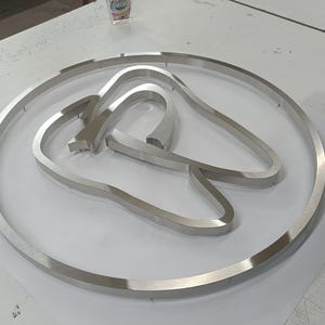 May include: A circular silver-toned metal sign featuring a stylized tooth outline centered within the frame. The metallic surface has a brushed finish, and the design consists of a continuous outer ring enclosing the tooth shape, creating a dimensional look.