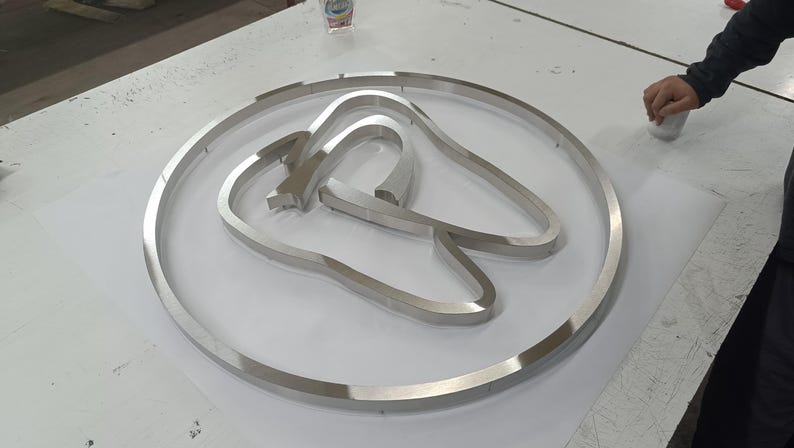 May include: A circular silver-toned metal sign featuring a stylized tooth outline centered within the frame. The metallic surface has a brushed finish, and the design consists of a continuous outer ring enclosing the tooth shape, creating a dimensional look.