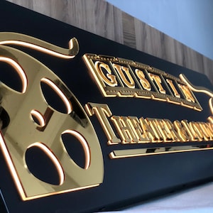 May include: A black sign with gold lettering and a gold film reel design. The sign reads "GUSTIN THEATER & LOUNGE".