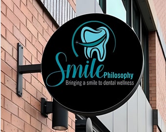 May include: A round black sign for a dental practice, mounted on a brick wall. The sign features the words "Smile Philosophy" in teal script, with a teal tooth graphic. The tagline reads "Bringing a smile to dental wellness."