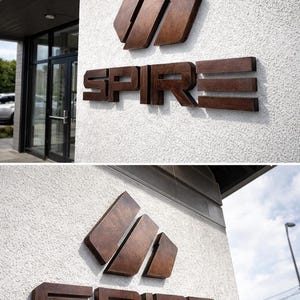 Puede incluir: Un letrero tridimensional montado en una pared blanca texturizada que presenta la palabra SPIRE en letras de bloque de color marr&oacute;n oscuro con un logotipo geom&eacute;trico a juego colocado directamente sobre el texto, creando una exhibici&oacute;n arquitect&oacute;nica.