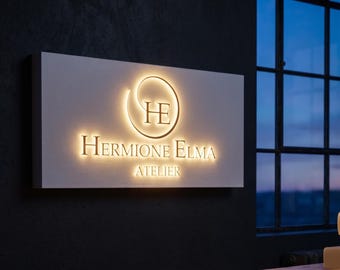 May include: Illuminated sign for "Hermione Elma Atelier" with a white rectangular base. The logo features a circular design with the letters "HE" and the name in a serif font. The word "ATELIER" is below the name.