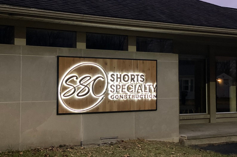 May include: A lit sign with a wooden background and black frame. The sign reads "SSC Shorts Specialty Construction" with a stylised logo of the letters "SSC" in a circle.
