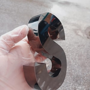 K&ouml;nnte beinhalten: Ein gl&auml;nzender, metallischer Buchstabe "S" mit reflektierender Oberfl&auml;che. Der Buchstabe hat eine dunkle, neutrale Farbe und eine dreidimensionale Erscheinung. Das Bild ist aus der Nahperspektive aufgenommen.