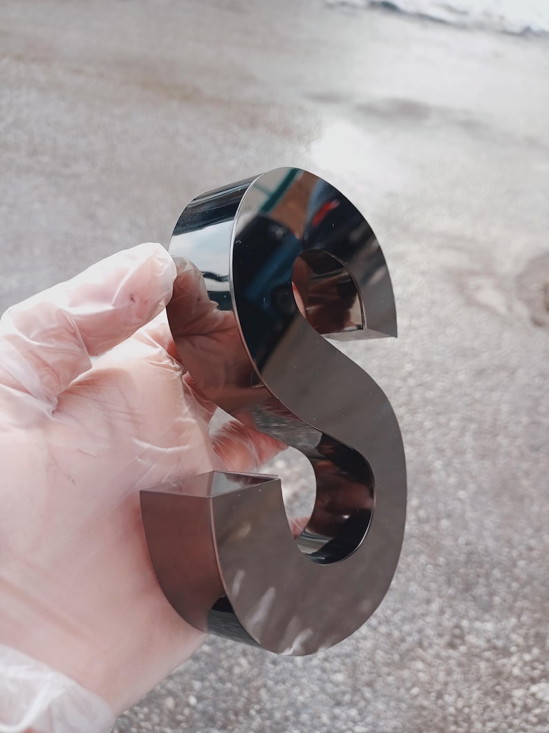 K&ouml;nnte beinhalten: Ein gl&auml;nzender, metallischer Buchstabe "S" mit reflektierender Oberfl&auml;che. Der Buchstabe hat eine dunkle, neutrale Farbe und eine dreidimensionale Erscheinung. Das Bild ist aus der Nahperspektive aufgenommen.