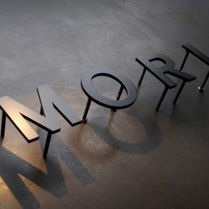 May include: Black metal letters spelling the word "MORE" are arranged on a dark surface. The letters cast long shadows, creating a dramatic effect. The letters are attached to the surface with small metal rods.