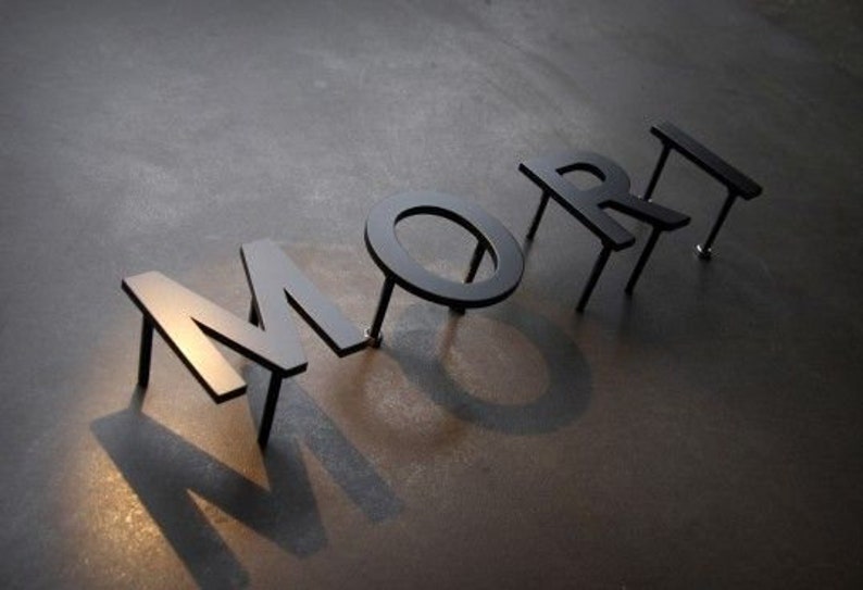 May include: Black metal letters spelling the word "MORE" are arranged on a dark surface. The letters cast long shadows, creating a dramatic effect. The letters are attached to the surface with small metal rods.