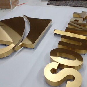 May include: A set of three-dimensional gold-colored metal signage letters spelling the word "Soulvet" alongside a stylized gold-colored triangular logo emblem. The letters and emblem feature a polished, reflective surface and raised edges.