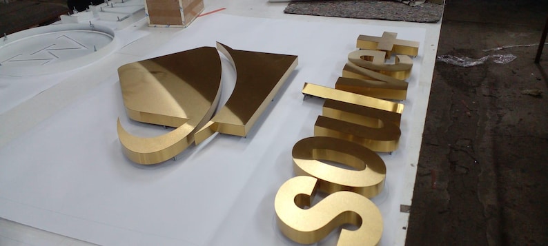 May include: A set of three-dimensional gold-colored metal signage letters spelling the word "Soulvet" alongside a stylized gold-colored triangular logo emblem. The letters and emblem feature a polished, reflective surface and raised edges.