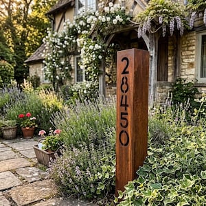 Puede incluir: Un poste de dirección rectangular y desgastado con el número "28450" recortado en negro. El poste, de metal marrón rojizo, se encuentra en un jardín con vegetación exuberante y un camino de piedra. Una casa de campo con encanto al fondo.