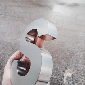 K&ouml;nnte beinhalten: Ein gro&szlig;er, gl&auml;nzender, silberner Metallbuchstabe "S" wird in einer behandschuhten Hand gehalten. Der Buchstabe hat ein abgerundetes, kursives Design mit flacher Ober- und Unterseite. Der Hintergrund ist eine verschwommene, neutral get&ouml;nte Au&szlig;enszene.