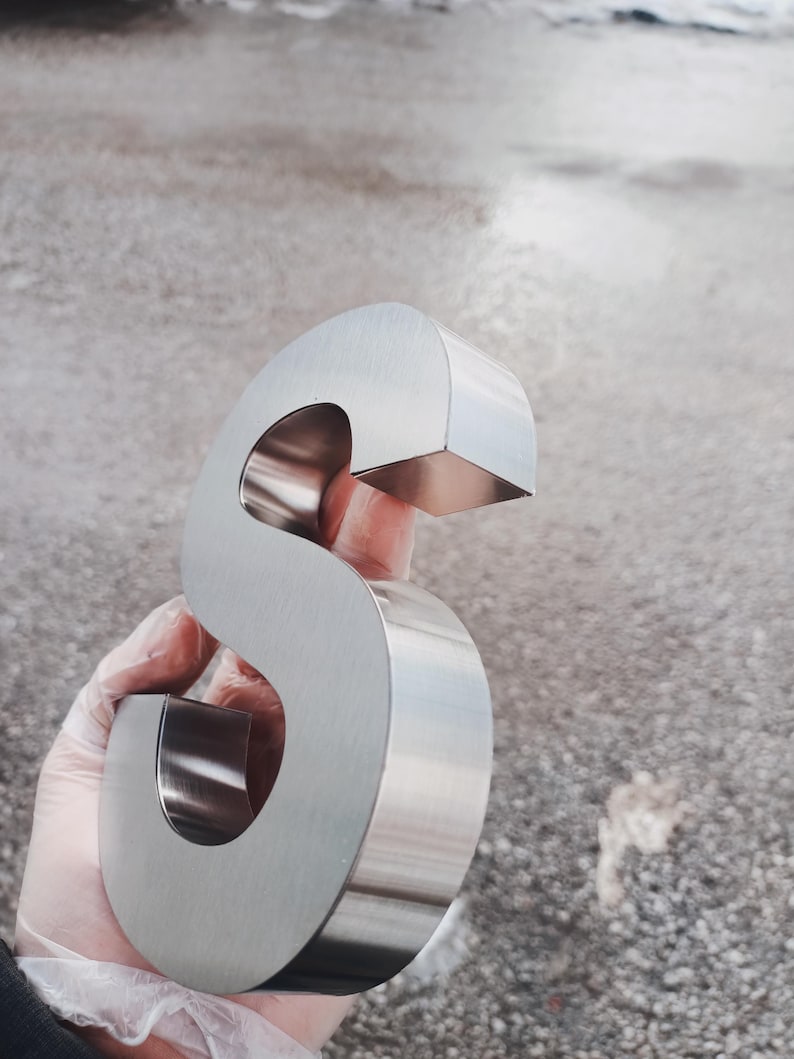 K&ouml;nnte beinhalten: Ein gro&szlig;er, gl&auml;nzender, silberner Metallbuchstabe "S" wird in einer behandschuhten Hand gehalten. Der Buchstabe hat ein abgerundetes, kursives Design mit flacher Ober- und Unterseite. Der Hintergrund ist eine verschwommene, neutral get&ouml;nte Au&szlig;enszene.