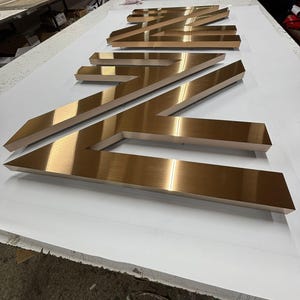 May include: Large, three-dimensional metal letters with a brushed bronze finish arranged in a row. The letters feature sharp, angular edges and a reflective surface, creating a bold, industrial appearance suitable for signage or architectural display.