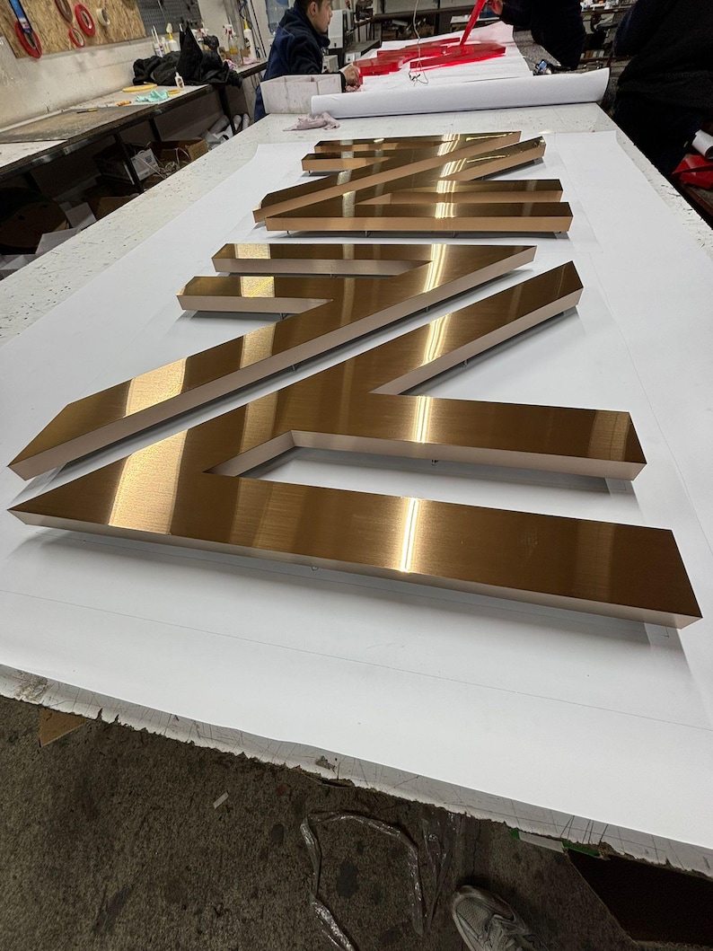 May include: Large, three-dimensional metal letters with a brushed bronze finish arranged in a row. The letters feature sharp, angular edges and a reflective surface, creating a bold, industrial appearance suitable for signage or architectural display.