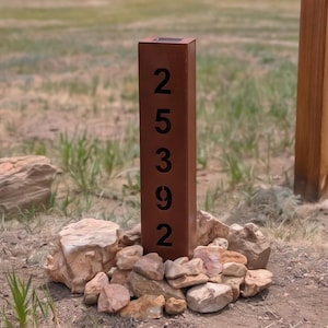 Può includere: Un alto palo rettangolare per numeri civici in metallo color ruggine, con i numeri neri 2, 5, 3, 9 e 2 allineati verticalmente. Il palo ha una sommità piatta ed è circondato alla base da un ammasso di rocce naturali di colore beige.
