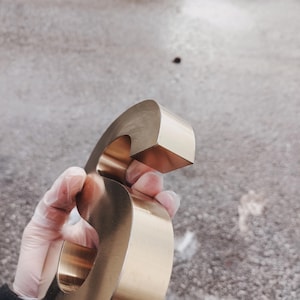 K&ouml;nnte beinhalten: Ein gro&szlig;er, metallisch goldener Buchstabe "G" wird in einer behandschuhten Hand gehalten. Der Buchstabe hat ein abgerundetes, blockartiges Design mit einer glatten, reflektierenden Oberfl&auml;che. Der Hintergrund ist eine strukturierte, graue Oberfl&auml;che.