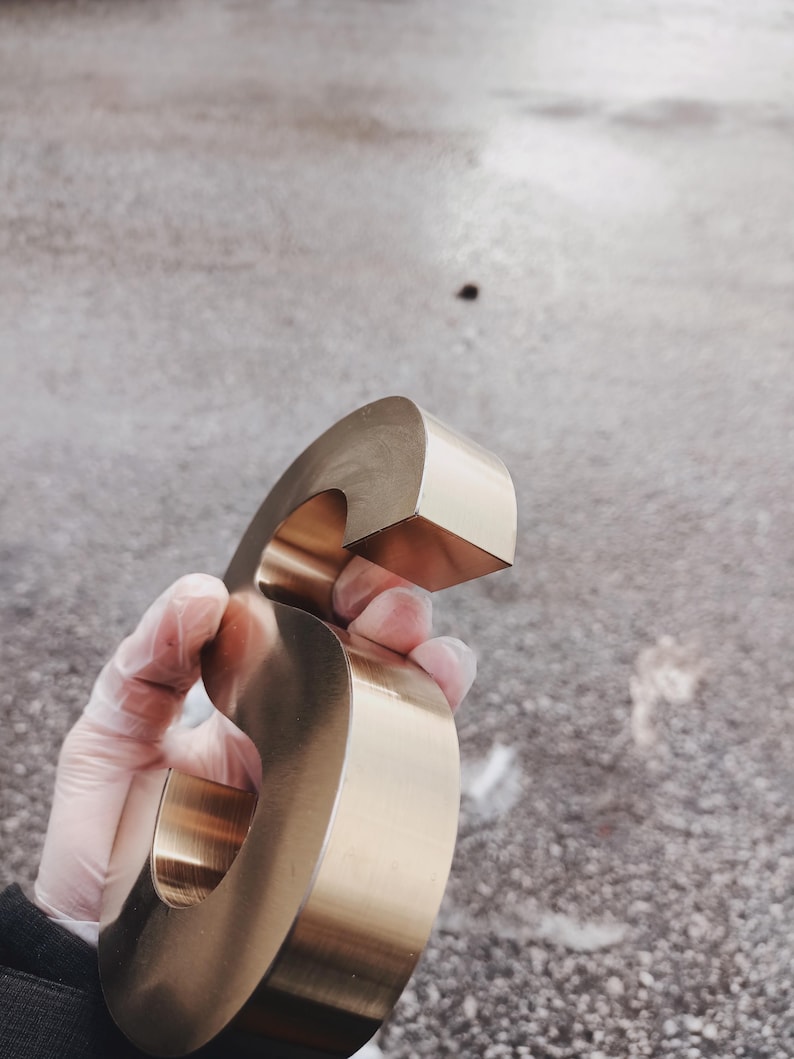 K&ouml;nnte beinhalten: Ein gro&szlig;er, metallisch goldener Buchstabe "G" wird in einer behandschuhten Hand gehalten. Der Buchstabe hat ein abgerundetes, blockartiges Design mit einer glatten, reflektierenden Oberfl&auml;che. Der Hintergrund ist eine strukturierte, graue Oberfl&auml;che.