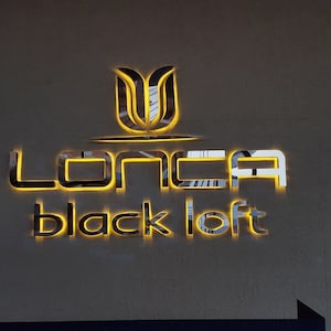 May include: A gold-coloured sign with the text "LONCA black loft" in a modern font. The sign is illuminated with warm yellow lights.
