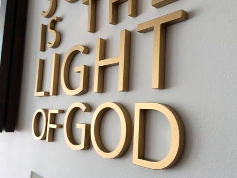 K&ouml;nnte beinhalten: Goldfarbene, dreidimensionale Buchstaben, die "SO THE LIGHT OF GOD" buchstabieren, sind an einer hellgrauen Wand angebracht. Die Buchstaben sind dick und werfen einen leichten Schatten, wodurch ein 3D-Effekt entsteht. Die &Auml;sthetik ist modern und inspirierend.