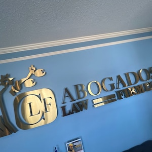 K&ouml;nnte beinhalten: Goldfarbenes Metallschild an einer blauen Wand. Das Schild zeigt die Buchstaben "LF" in einem Quadrat und die Worte "ABOGADOS LAW FIRM" in Gro&szlig;buchstaben. Eine stilisierte Figur einer Person ist ebenfalls Teil des Schildes.