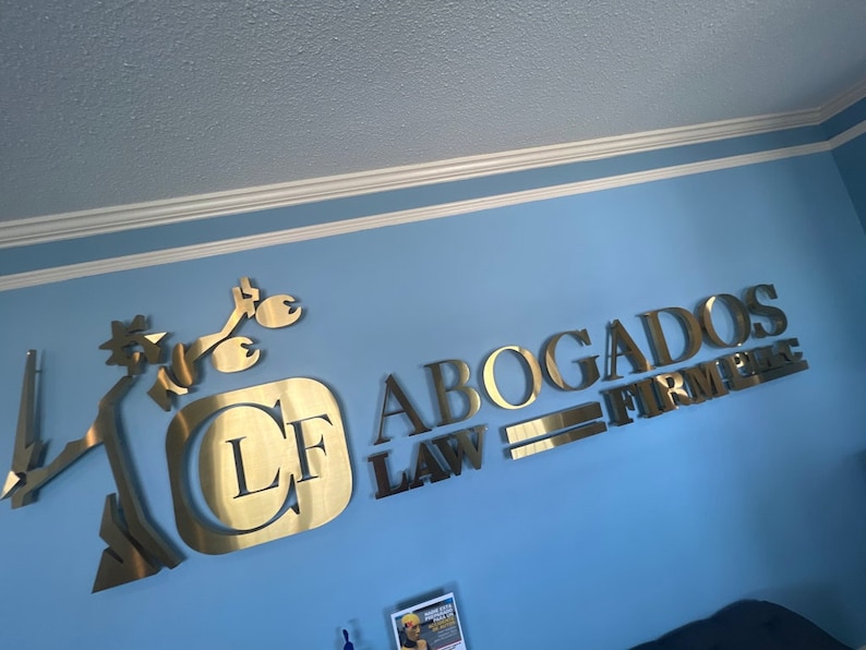 K&ouml;nnte beinhalten: Goldfarbenes Metallschild an einer blauen Wand. Das Schild zeigt die Buchstaben "LF" in einem Quadrat und die Worte "ABOGADOS LAW FIRM" in Gro&szlig;buchstaben. Eine stilisierte Figur einer Person ist ebenfalls Teil des Schildes.