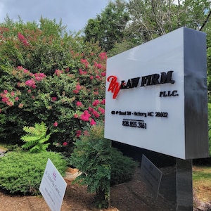 Puede incluir: Un letrero rectangular blanco para exteriores de RV Law Firm PLLC con letras negras y un logotipo rojo. El letrero muestra la direcci&oacute;n 409 4th Street SW - Hickory, NC 28602 y el n&uacute;mero 828.855.3161. Se ve un letrero blanco m&aacute;s peque&ntilde;o al lado.