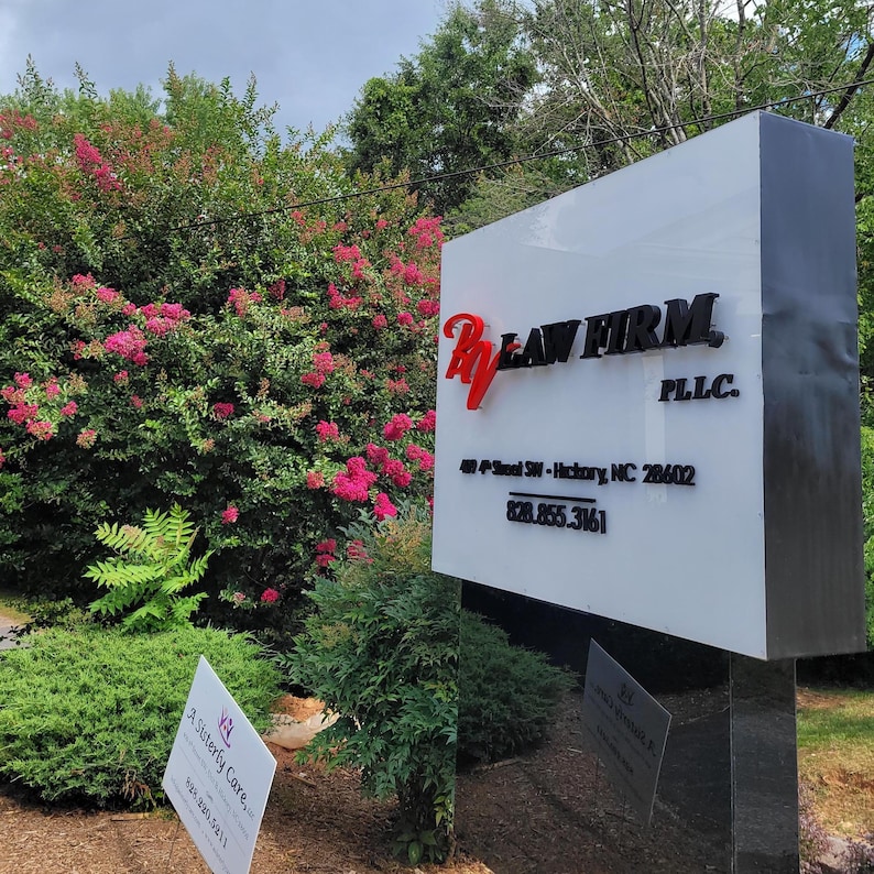 Puede incluir: Un letrero rectangular blanco para exteriores de RV Law Firm PLLC con letras negras y un logotipo rojo. El letrero muestra la direcci&oacute;n 409 4th Street SW - Hickory, NC 28602 y el n&uacute;mero 828.855.3161. Se ve un letrero blanco m&aacute;s peque&ntilde;o al lado.
