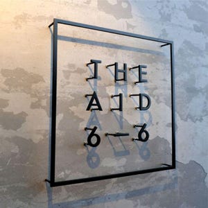 May include: Black metal wall art with the words "THE AID 6-6" mounted on a clear panel. The letters and numbers are in a bold, sans-serif font. The artwork is framed by a black square and mounted on a textured gray wall.
