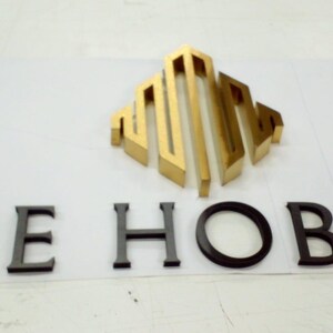 May include: A three-dimensional sign featuring a gold metallic geometric logo above the black text THE HOBART. The letters and logo are mounted on a flat white surface, creating a raised effect with clean, sharp edges and a professional finish.