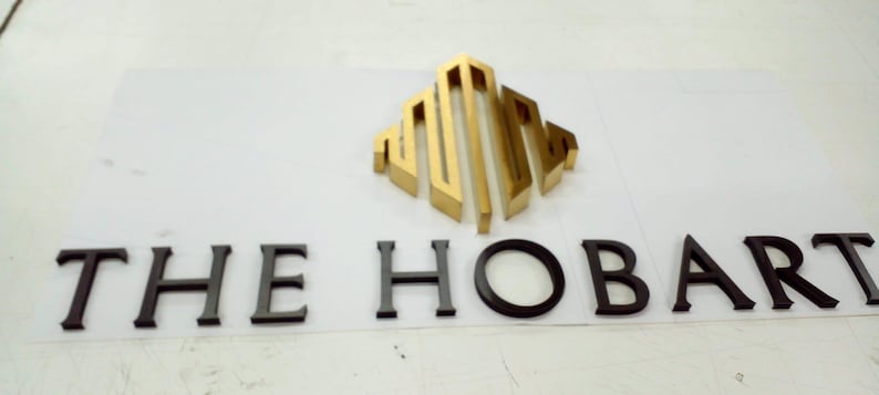 May include: A three-dimensional sign featuring a gold metallic geometric logo above the black text THE HOBART. The letters and logo are mounted on a flat white surface, creating a raised effect with clean, sharp edges and a professional finish.