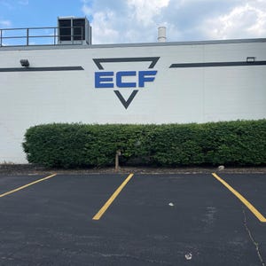 Puede incluir: Una pared exterior blanca de un edificio con un gran logotipo triangular negro con las letras azules ECF centradas en su interior. El edificio tiene una franja horizontal oscura, una unidad de climatizaci&oacute;n en el techo y un seto verde.