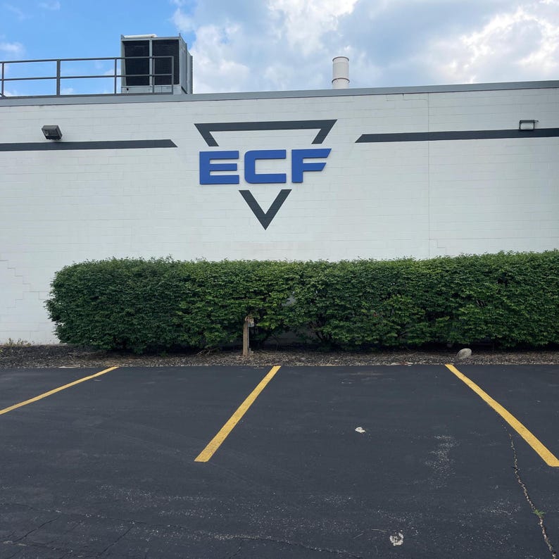Puede incluir: Una pared exterior blanca de un edificio con un gran logotipo triangular negro con las letras azules ECF centradas en su interior. El edificio tiene una franja horizontal oscura, una unidad de climatizaci&oacute;n en el techo y un seto verde.