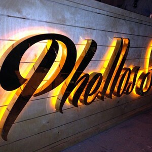 May include: A black illuminated sign with the word "Phellad" in a stylized font, mounted on a wood panel background.