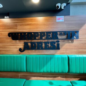 May include: A restaurant interior featuring a wooden wall with the words "Whopper in Adresi" in black lettering. Below, a green cushioned bench is visible. Security cameras are mounted on the ceiling.
