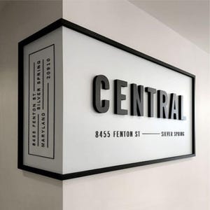 May include: Black and white sign with a black frame and the word "CENTRAL" in large, bold, black letters. The address "8455 Fenton St - Silver Spring" is printed below the word "CENTRAL". The address "8455 Fenton St Silver Spring 20910 Maryland" is printed on the left side of the sign.