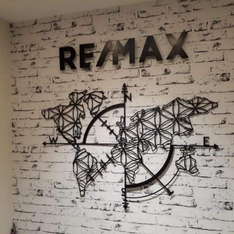 May include: Black metal wall art featuring the word "RE/MAX" above a world map design. The map is a stylized representation with compass details. The artwork is mounted on a white brick wall.