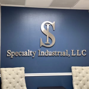 May include: A large, silver-colored metal sign mounted on a dark blue wall. The sign features a stylized letter S with an I integrated into its center, positioned above the text Specialty Industrial, LLC in a clean, sans-serif font.