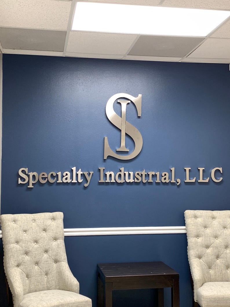 May include: A large, silver-colored metal sign mounted on a dark blue wall. The sign features a stylized letter S with an I integrated into its center, positioned above the text Specialty Industrial, LLC in a clean, sans-serif font.