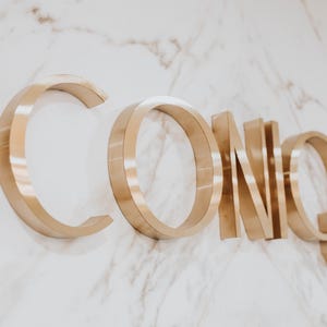 May include: Three-dimensional metallic gold lettering spelling the word ICONIQUE mounted on a white marble surface with grey veining. The letters have a polished finish and cast subtle shadows, creating a depth effect against the stone background.