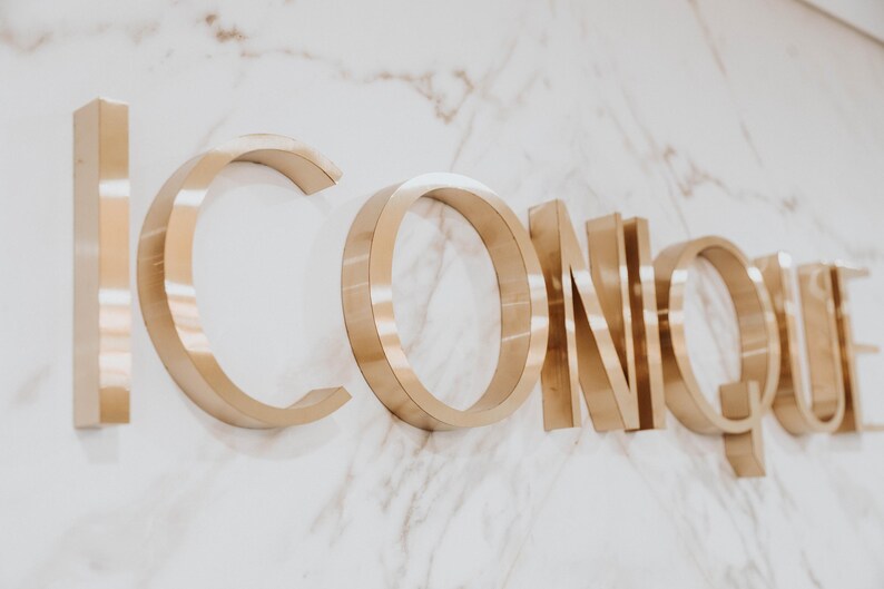 May include: Three-dimensional metallic gold lettering spelling the word ICONIQUE mounted on a white marble surface with grey veining. The letters have a polished finish and cast subtle shadows, creating a depth effect against the stone background.