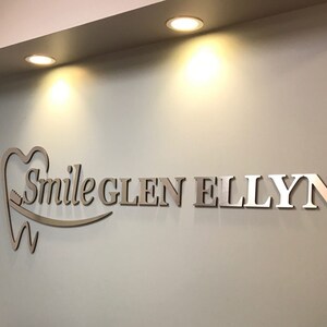 May include: A three-dimensional wall sign featuring the text Smile GLEN ELLYN in a metallic finish. The logo includes a stylized tooth icon with a toothbrush integrated into the design, mounted on a light-colored wall under recessed lighting.