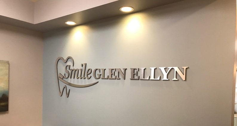 May include: A three-dimensional wall sign featuring the text Smile GLEN ELLYN in a metallic finish. The logo includes a stylized tooth icon with a toothbrush integrated into the design, mounted on a light-colored wall under recessed lighting.