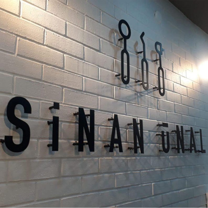 May include: Black metal sign on a white brick wall. The sign reads "SINAN UNAL" in bold, capital letters. Above the name are three decorative black metal shapes. The wall is lit by a warm light.