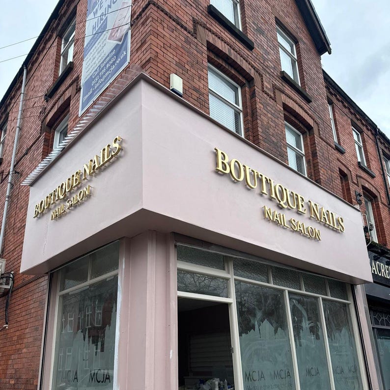 Puede incluir: Un local comercial en esquina con fachada rosa claro y letras doradas que dicen BOUTIQUE NAILS NAIL SALON. El edificio es de ladrillo rojo con varias ventanas de marco blanco y una gran entrada acristalada en la planta baja.
