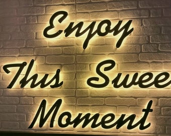 May include: A white brick wall with a black neon sign that reads "Enjoy This Sweet Moment".