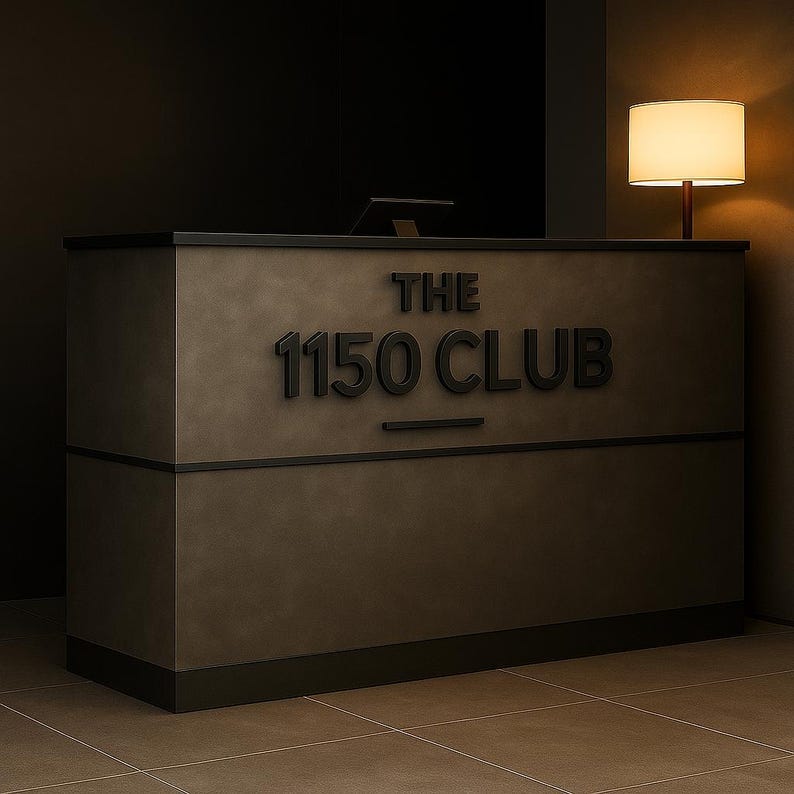 May include: A rectangular reception desk with a matte dark gray finish and a black base. The front features raised black lettering that reads THE 1150 CLUB with a horizontal line underneath. A small lamp with a warm white shade sits to the right.