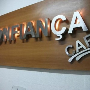 May include: A wooden sign with the words "CONFIAN&Ccedil;A CAR" in raised, silver letters. The letters are illuminated from behind, creating a warm glow.